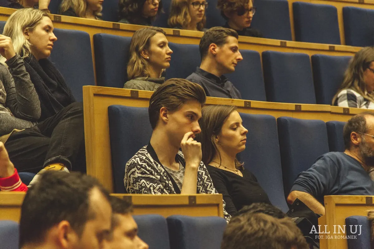 An audience in blue seats in a lecture hall, with two people in the centre listening closely.