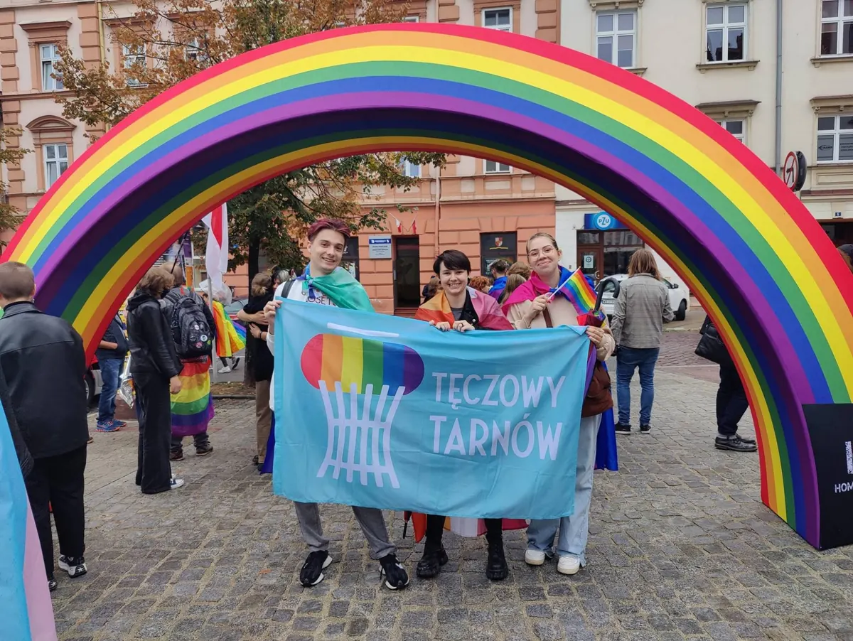 Three people holding the Tęczowy Tarnów flag under a rainbow arch in a town square.