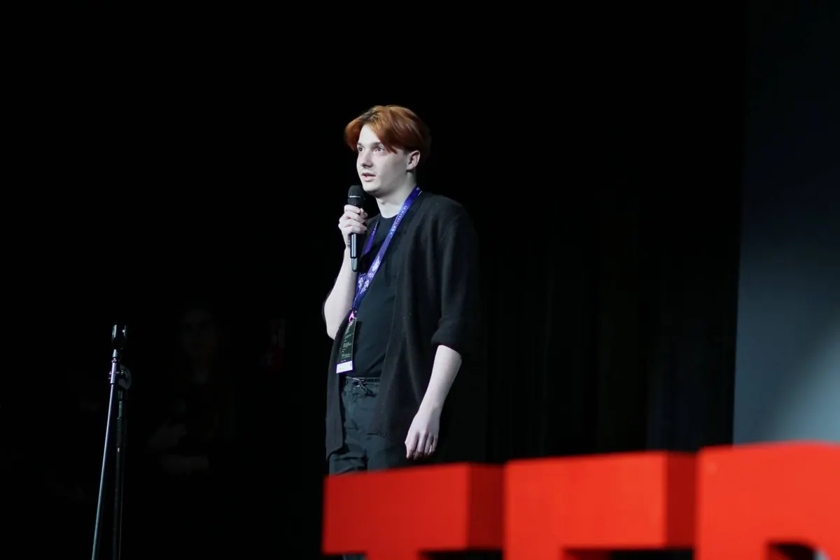 Artur Sendyka holding a microphone on a dark stage, with red decorative letters in the foreground.